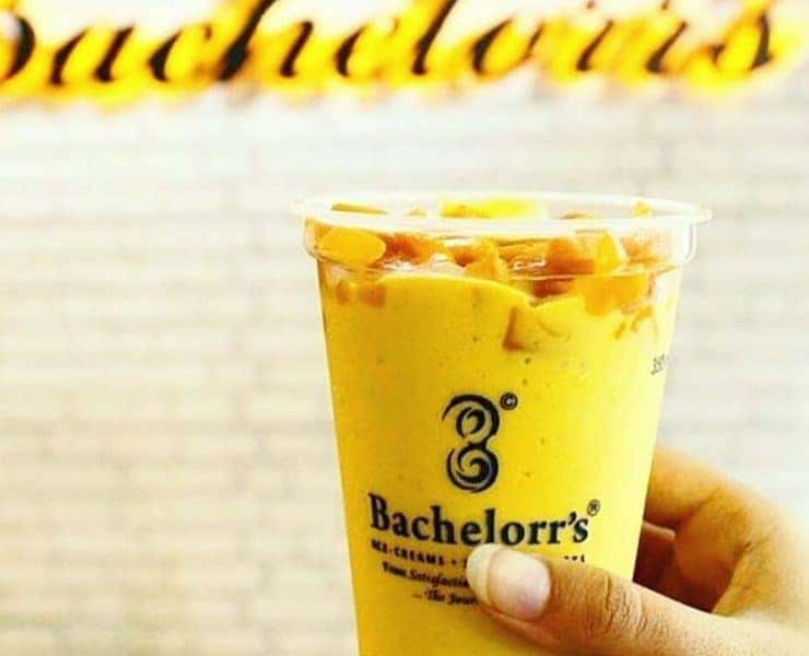 A person holding up a cup of orange juice in front of a sign that says bachelor's.
