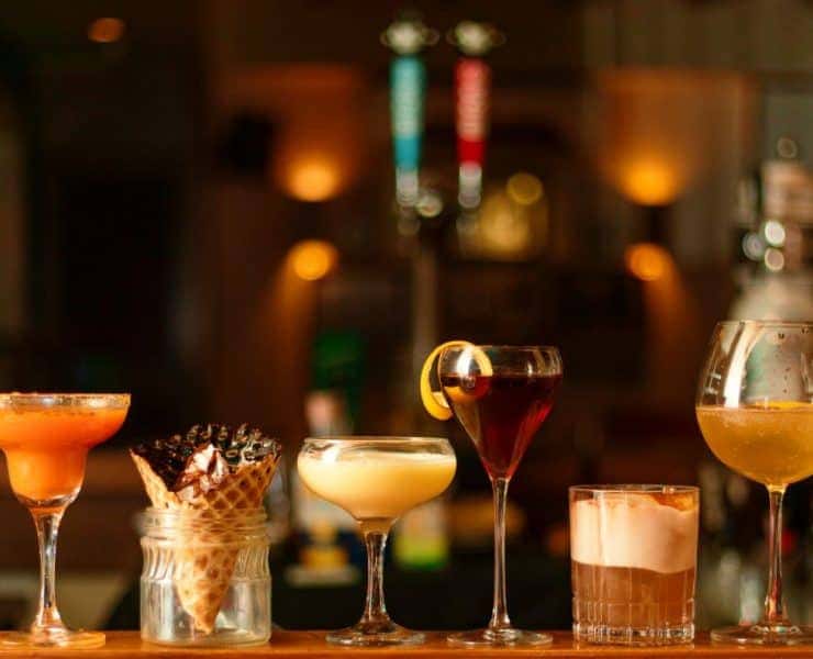 A row of five cocktails on a bar counter, each in a different glass, with various garnishes under warm ambient lighting.