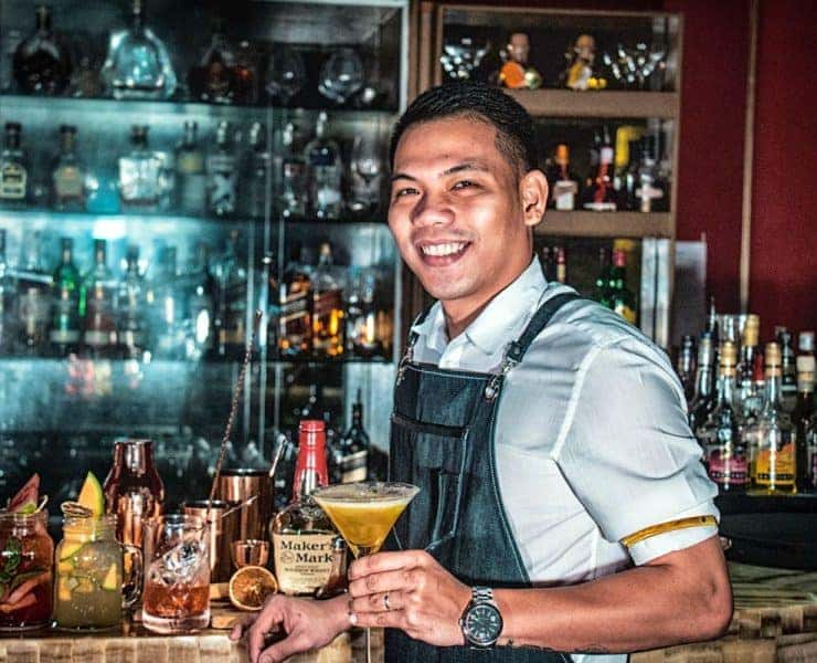 Bartender holding a cocktail, standing in front of a bar with various bottles, jars, and a Maker's Mark bourbon bottle visible.