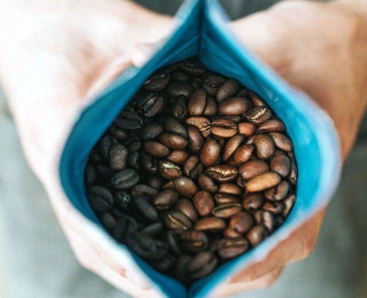 Hands holding an open blue bag filled with roasted coffee beans.