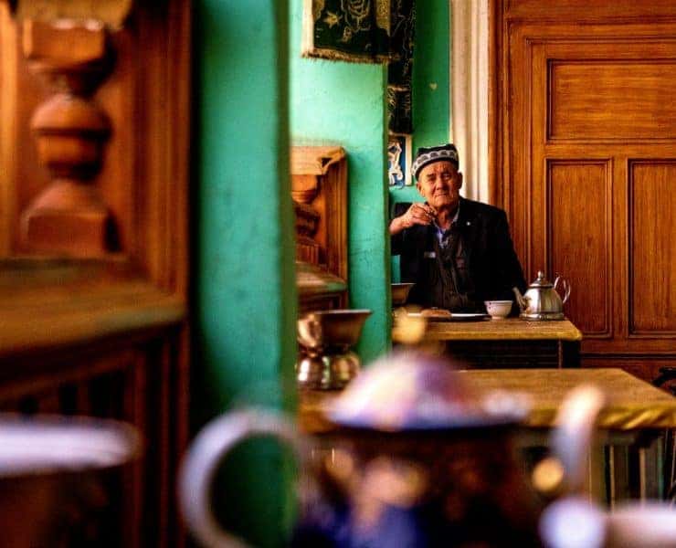 A person wearing a patterned cap sits at a table in a room with green walls and wooden details, holding a cup and looking into the distance.