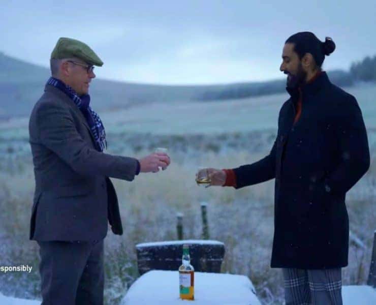 Two men stand outdoors in a snowy landscape, holding glasses of whiskey. A bottle is on the table between them.