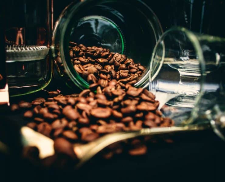 Coffee beans spilling from a tipped glass jar onto a dark surface.