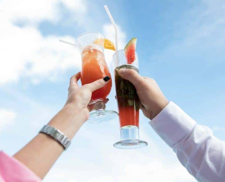 Two people holding glasses with drinks against a blue sky, each with a straw and garnished with fruit slices.