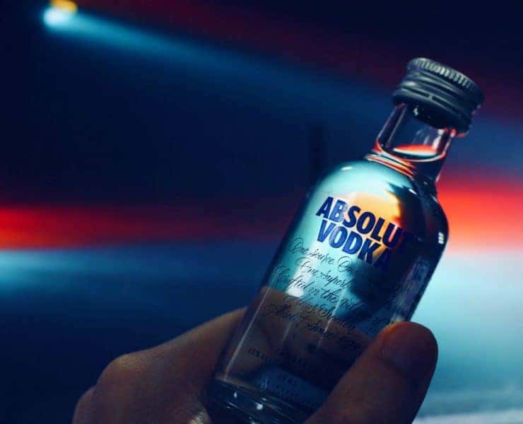 Hand holding a small bottle of Absolut Vodka against a colorful, blurred background.