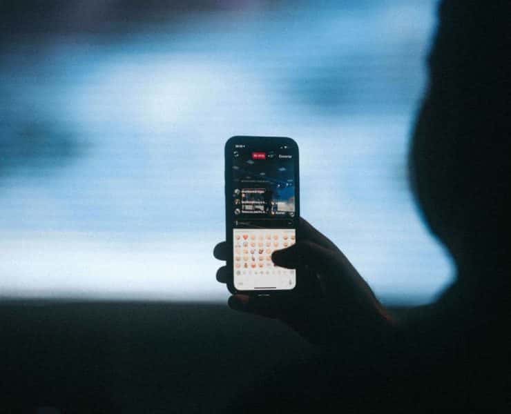 A hand holding a smartphone with a live streaming video on the screen, typing with a visible keyboard. The background is blurred.
