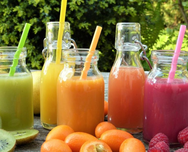 Four glass bottles with colorful fruit smoothies and straws, surrounded by kiwi, kumquats, raspberries, and grapefruit, on a wooden table outdoors.