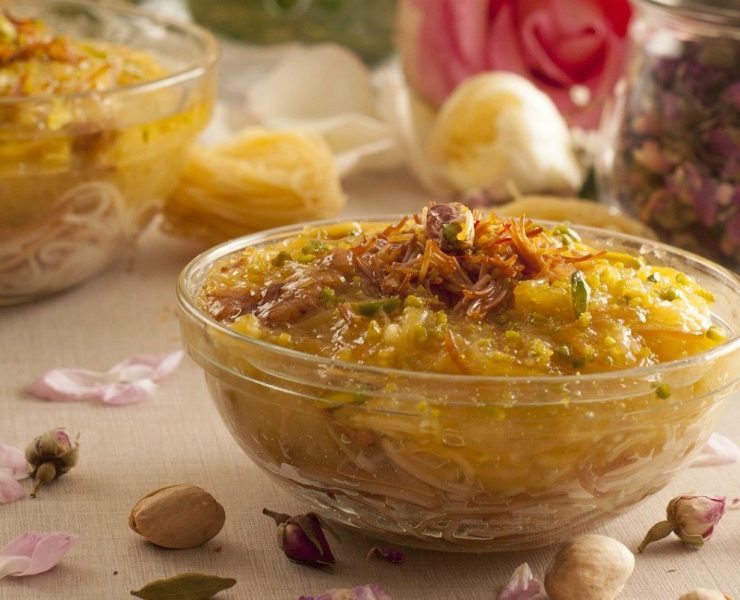 Glass bowls filled with a saffron-colored dessert topped with nuts, surrounded by scattered petals and nuts on a cream-colored cloth. A rose and jars sit in the background.