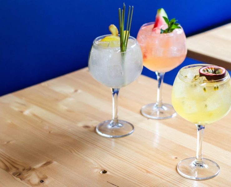 Three colorful cocktails in goblet glasses are arranged on a wooden table against a blue background. Each drink is garnished with different fruits and herbs.