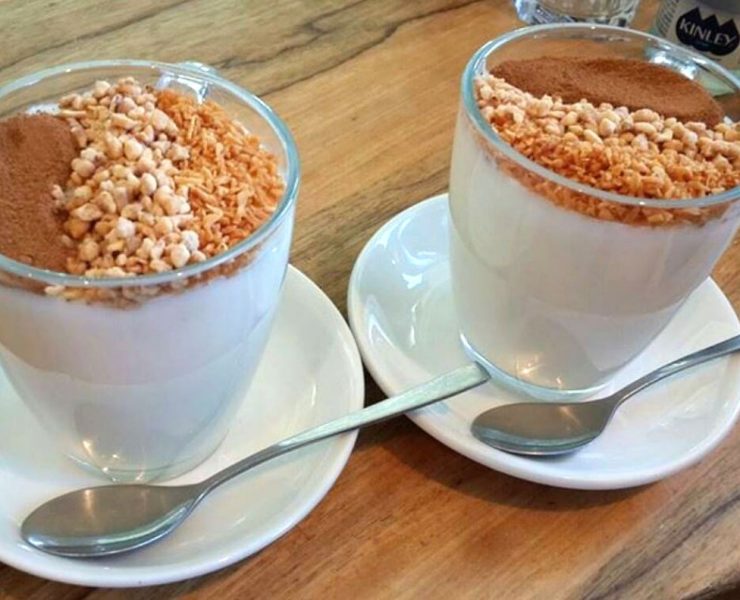 Two glasses of creamy dessert topped with a layer of cereal, cocoa powder, and toasted coconut flakes, served on white saucers with spoons.