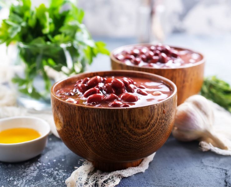 Two wooden bowls filled with cooked red beans in sauce. In the background, there's a bunch of fresh parsley and a clove of garlic on a textured surface.