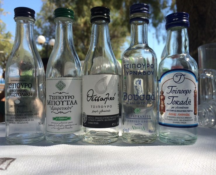 Five empty Tsipouro bottles with Greek labels are lined up on a table outdoors, with trees blurred in the background.