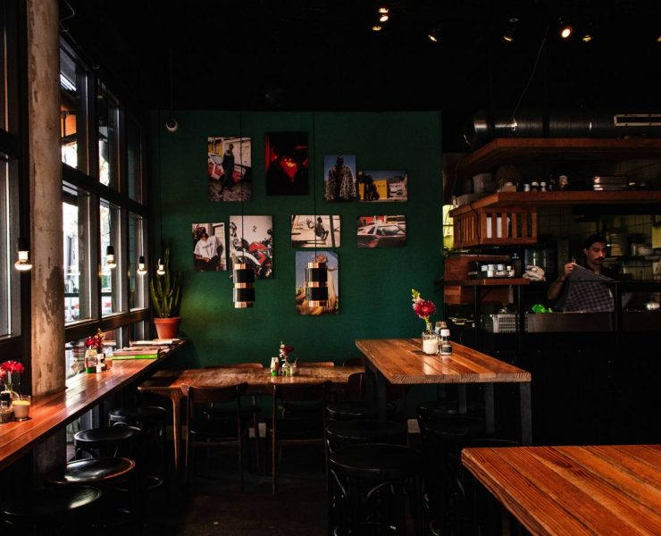 A cozy café with wooden tables, greenery, and framed photos on a dark green wall. A counter area with a person in the background is visible.