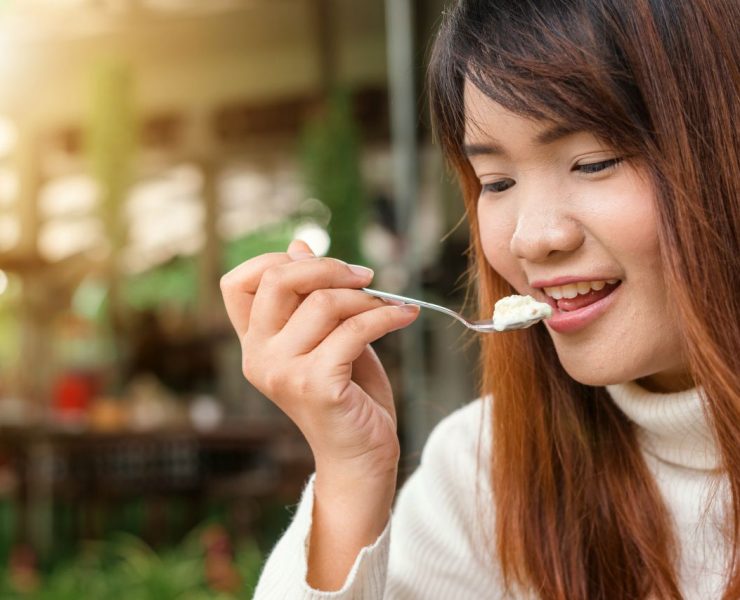 A woman with long hair smiles while holding a spoon near her mouth. She is wearing a white sweater, and the background appears to be a blurred outdoor setting.