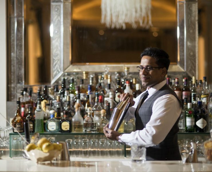 Bartender in a vest shaking a cocktail shaker behind a bar stocked with various bottles and a bowl of lemons.