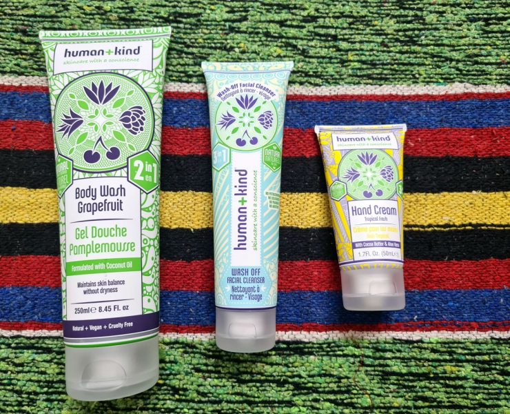 Three tubes of human+kind products, including a body wash, face cleanser, and hand cream, placed on a colorful woven fabric background.