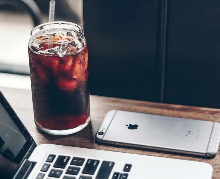 A glass of iced coffee with a straw next to an open laptop and a smartphone on a wooden table.