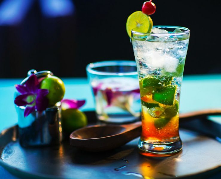 A tall glass of iced cocktail with lime and mint on a tray, accompanied by a shorter glass, fresh limes, and a purple flower.