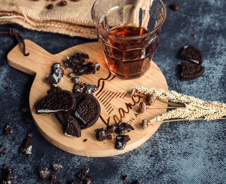 A glass of dark liquid on a wooden board with chocolate cookie pieces, surrounded by wheat stalks.