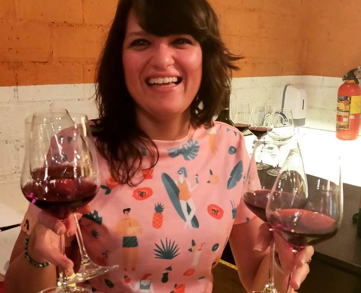 A person smiling, holding three glasses of red wine, in a room with orange and white walls.