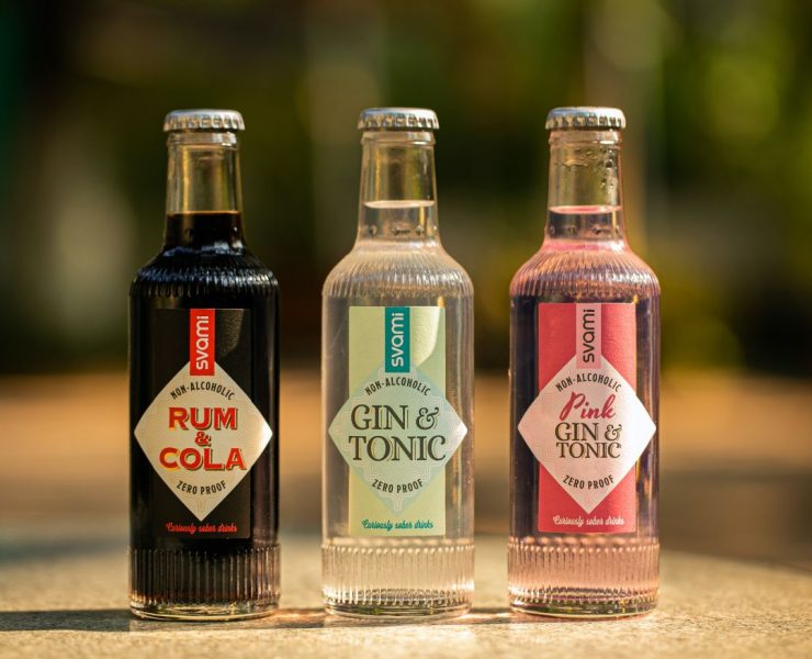 Three bottles of non-alcoholic drinks labeled Rum & Cola, Gin & Tonic, and Pink Gin & Tonic are displayed in a row on a sunlit surface.