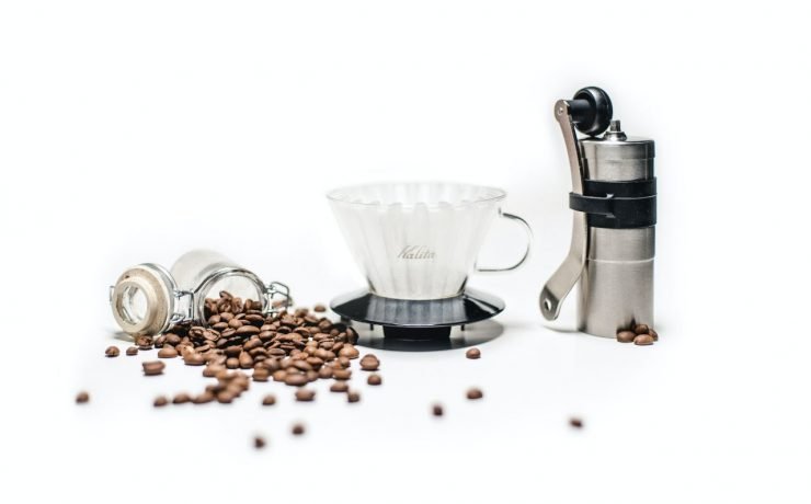 Coffee beans spill from a jar next to a glass coffee dripper and a hand coffee grinder on a white surface.