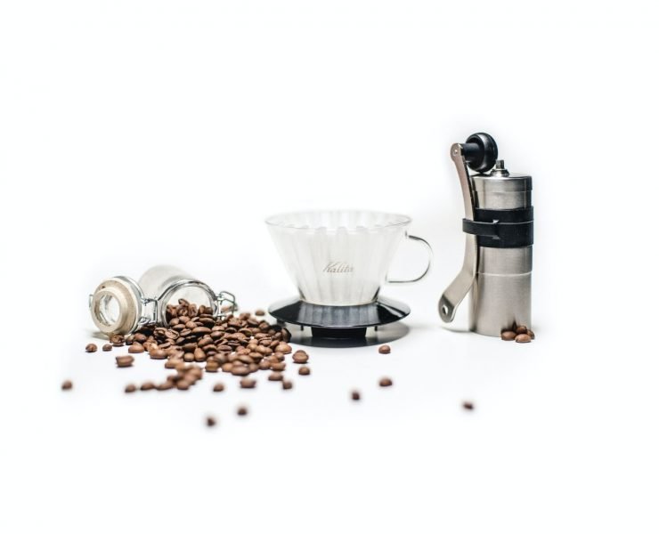 Coffee beans spill from a jar next to a glass coffee dripper and a hand coffee grinder on a white surface.