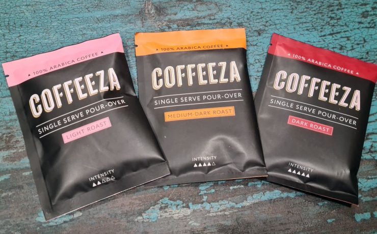 Three Coffeeza single-serve pour-over coffee packets displayed on a textured blue surface. Flavors: Light Roast, Medium-Dark Roast, Dark Roast.