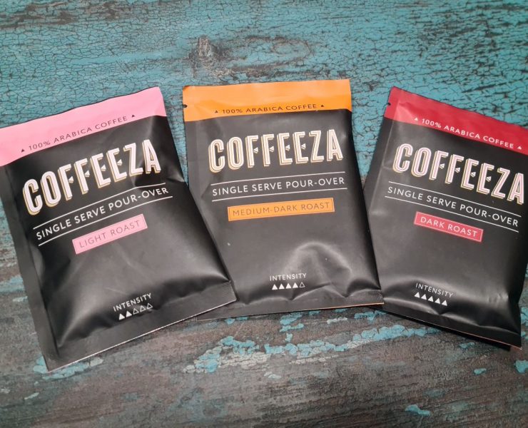 Three Coffeeza single-serve pour-over coffee packets displayed on a textured blue surface. Flavors: Light Roast, Medium-Dark Roast, Dark Roast.