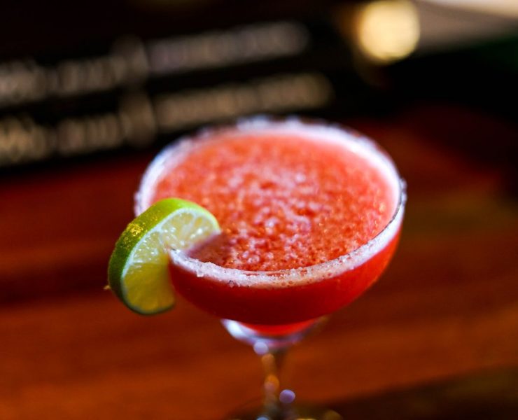 A red frozen cocktail in a glass garnished with a lime wedge, set on a wooden table.