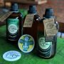 Three glass beer bottles with labels and tags are on a green table, accompanied by a coaster with "t WIT Belgian Wheat" text and a logo. A brick wall is in the background.