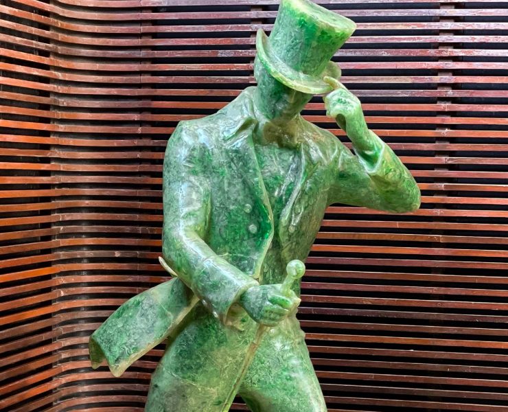 A green statue of a person wearing a top hat and holding a cane, posed in front of a wooden slatted background.