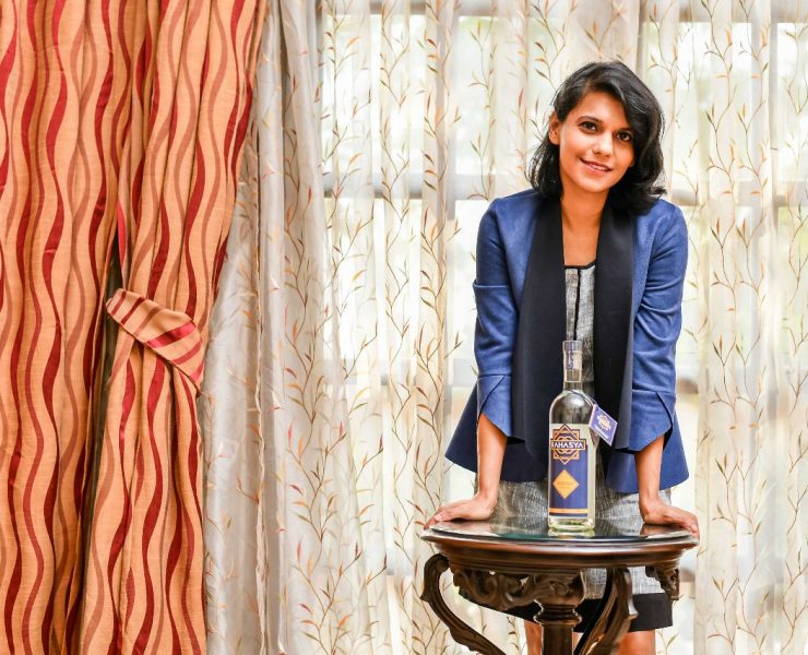Woman in a blue jacket stands behind a wooden table with a bottle on it, in front of patterned curtains.
