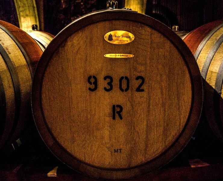 Close-up of a wooden barrel with the numbers "9302 R" painted on it. The barrel is part of a row in a dimly lit cellar.