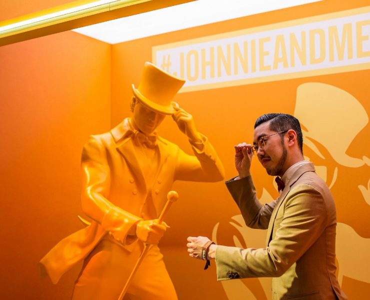 A man in a suit poses with an orange statue of a gentleman holding a cane and tipping his hat. Background text reads "#JOHNNIEANDME.