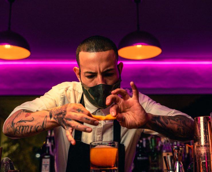 Bartender with tattoos garnishes a cocktail with an orange peel. He's focused, wearing a mask, under warm lighting in a bar setting.