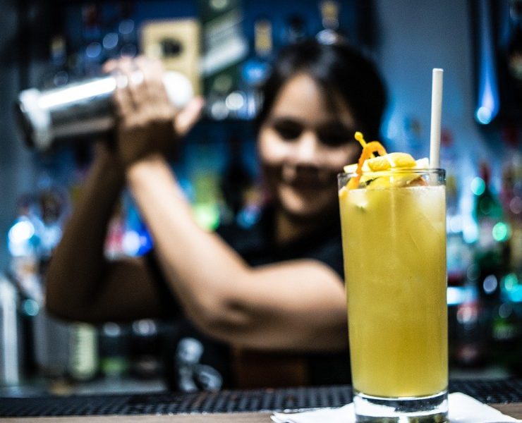 female bartender shaking cocktail in the background with a yellow cocktail in front