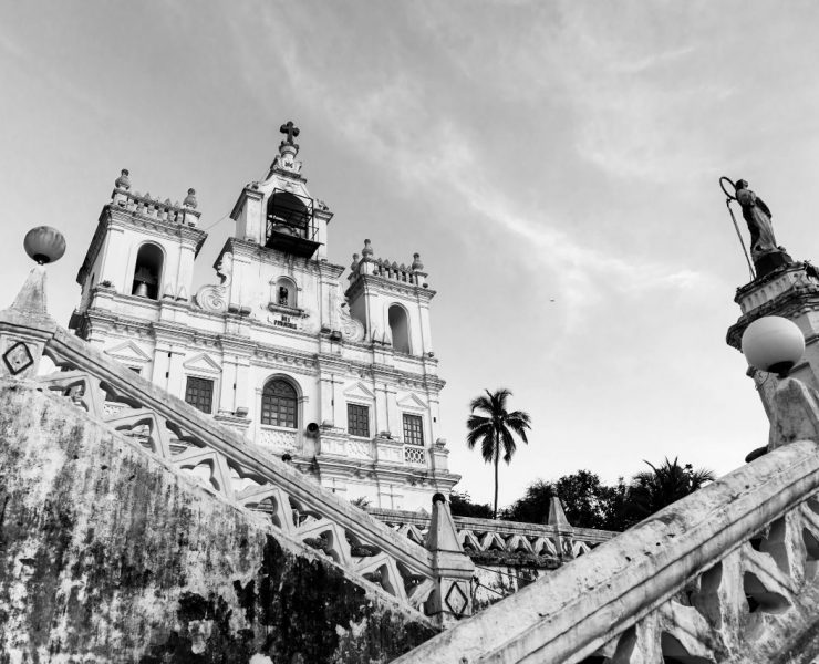 A black and white photo of a colonial-style church with ornate details and stairs leading up, surrounded by statues and a palm tree in the background.