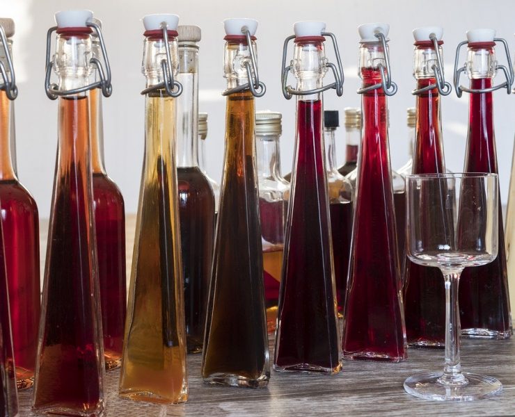 Several glass bottles filled with various colorful liquids are arranged on a wooden surface. A single empty glass stands in front of the bottles.