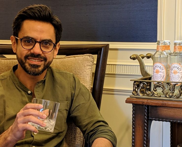 A person holding a glass with ice sits on a couch next to a table with bottles of drink in a tray.