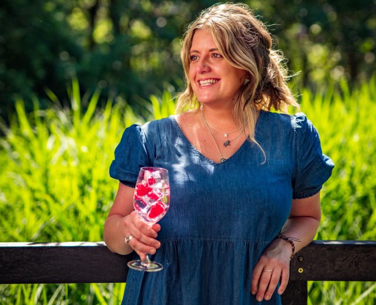 Woman in blue dress smiles and holds a glass with a pink drink and ice cubes outdoors, against a backdrop of greenery.