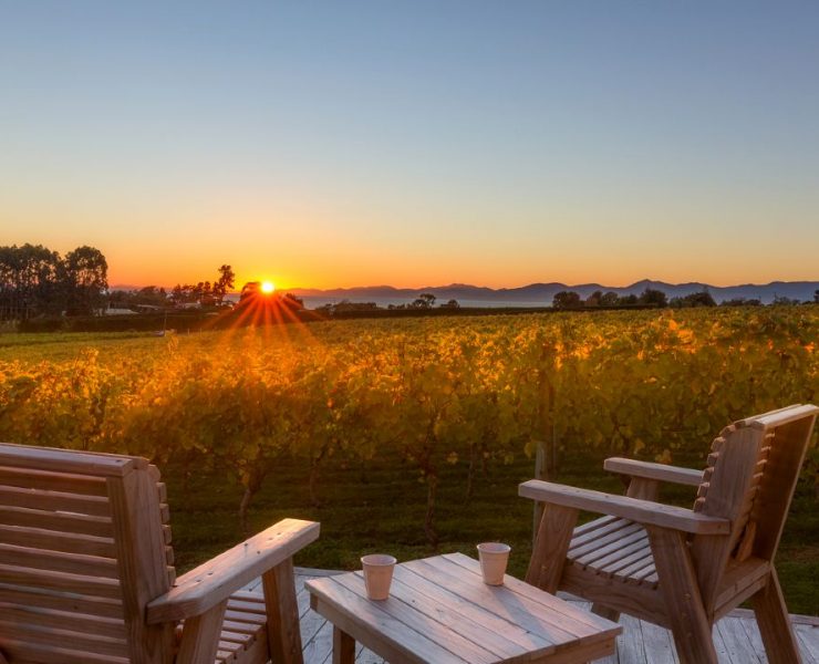 Two wooden chairs face a vineyard at sunset with mountains in the background. A small table holds two cups.