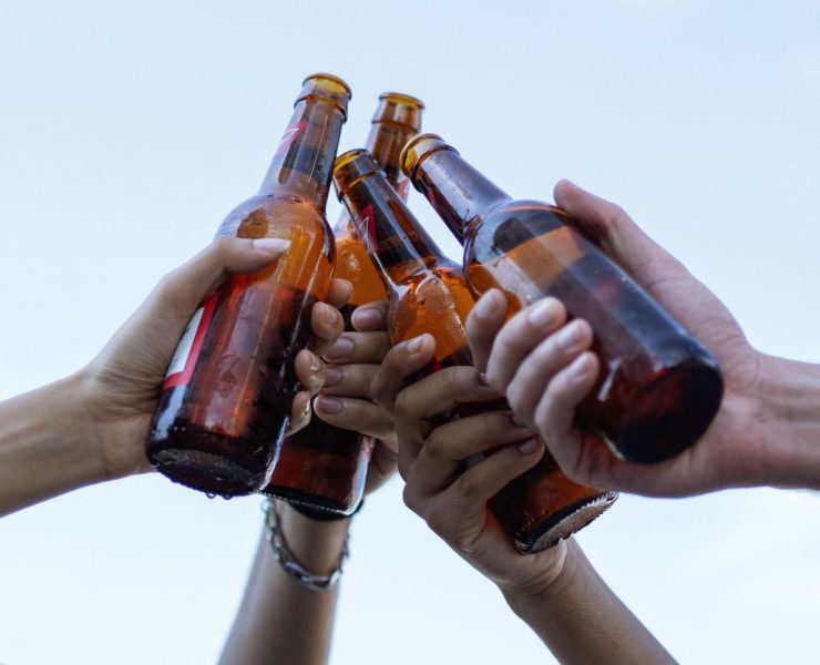 Five hands clinking brown glass beer bottles against a clear sky.