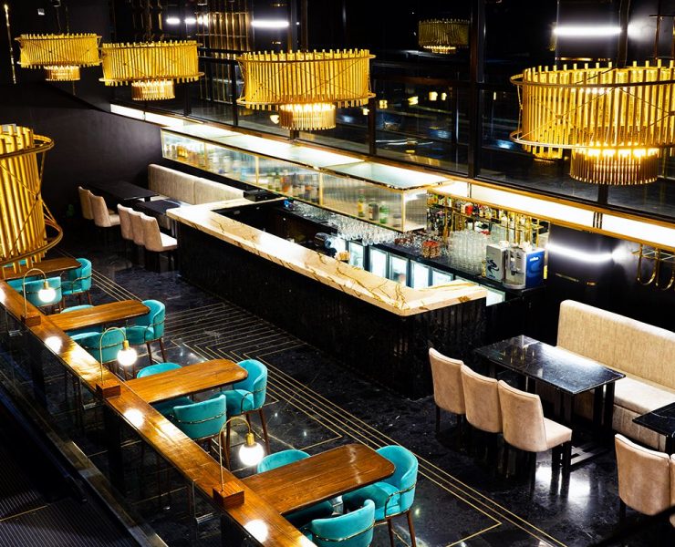 Elegant restaurant interior with gold lighting fixtures, polished black flooring, teal and beige seating, and a central bar with a marble counter.