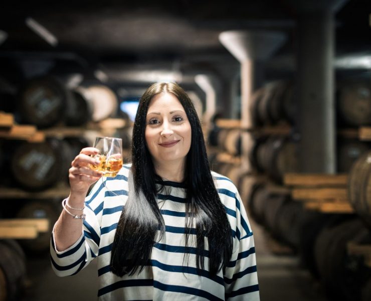 A person in a striped shirt holds a glass of whiskey, standing in a dimly lit barrel room.