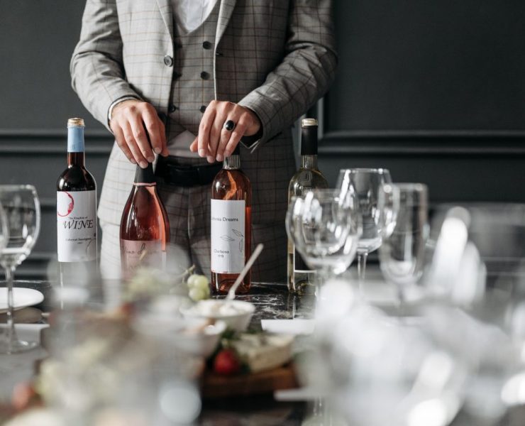 A person in a plaid suit is arranging three wine bottles on a table set with empty wine glasses and a charcuterie board.