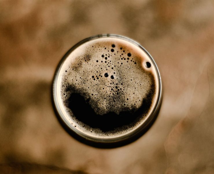 Top view of a glass of dark beer with a creamy head, set against a blurred brown background.