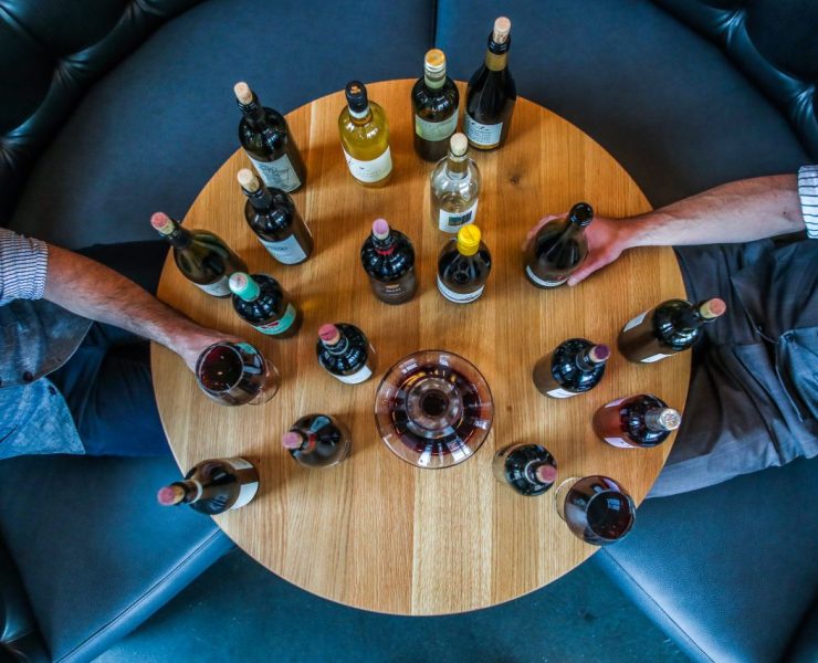 A round wooden table with various wine bottles and two people sitting opposite each other, each holding a bottle.
