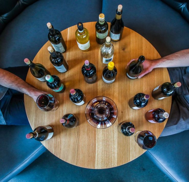 A round wooden table with various wine bottles and two people sitting opposite each other, each holding a bottle.