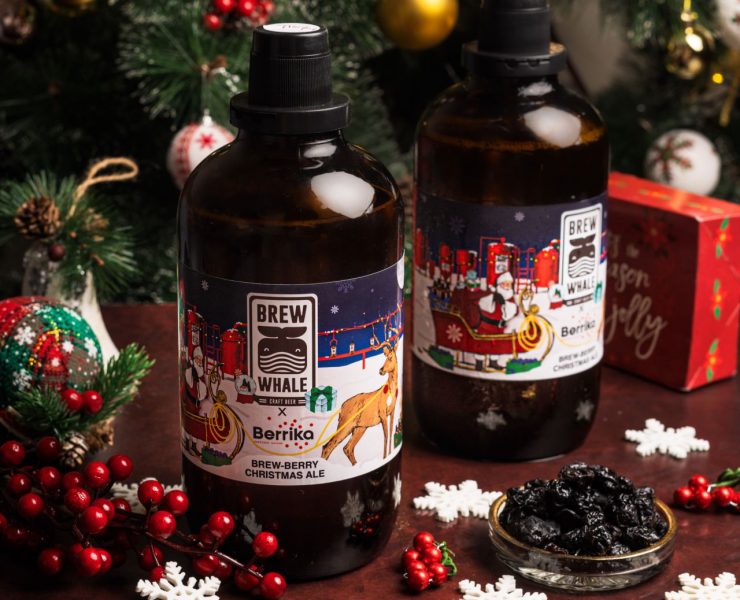 Two bottles of Brew Whale Beriiko Christmas Ale on a table with festive decorations, red berries, and a small bowl of dried fruit, in front of a decorated Christmas tree.